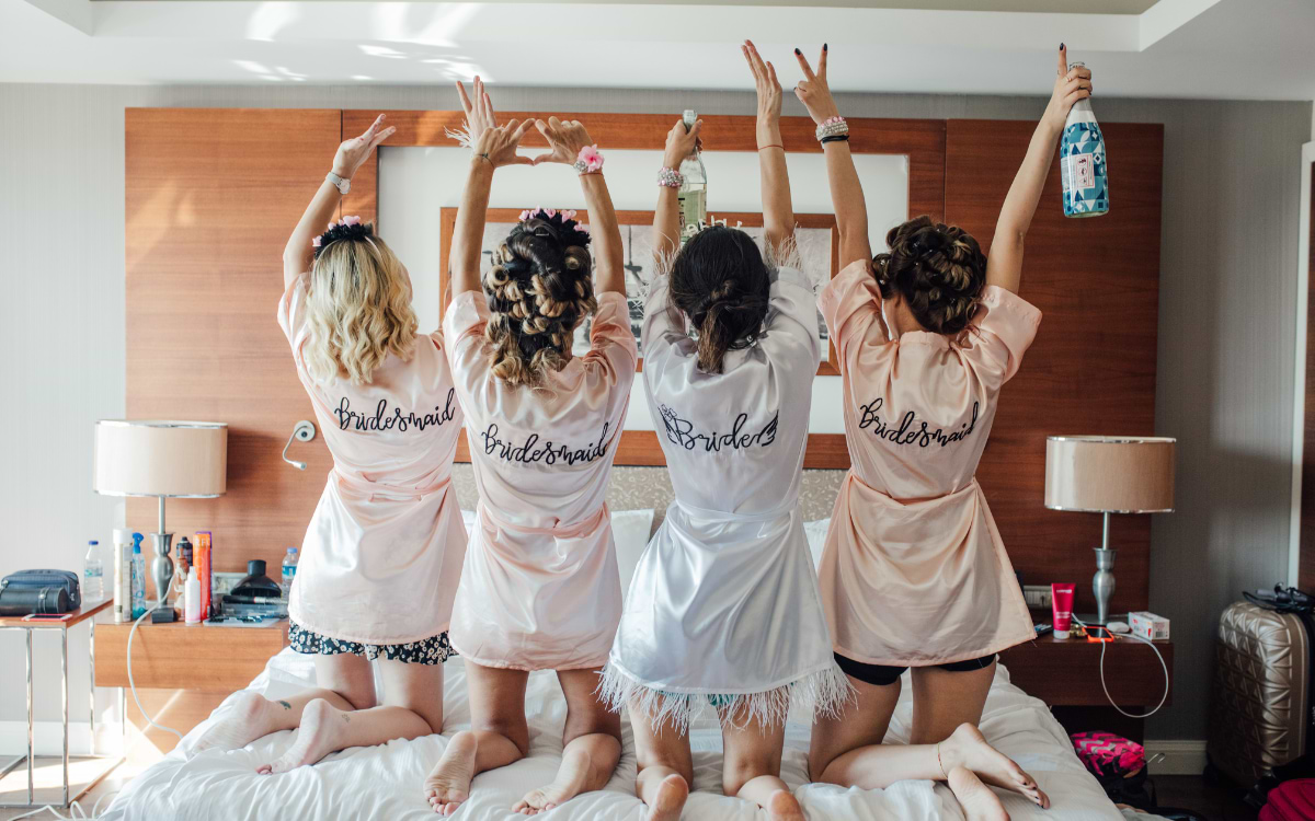 Four women in satin robes on a hotel bed celebrating the bride to be