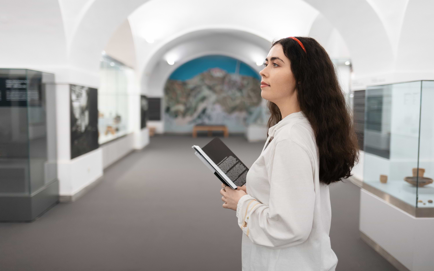 A young woman reads a museum guidebook inside a vaulted underground exhibition hall lined with glass display cases