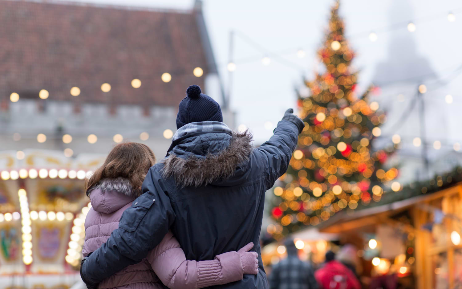Couple in winter coats embracing at a Christmas market, pointing towards a lit tree and a vintage carousel