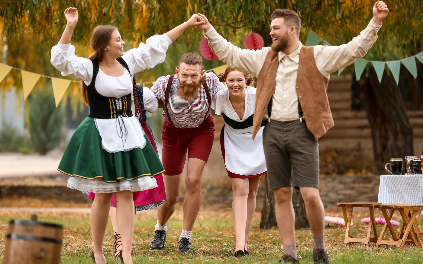 Friends in dirndls and lederhosen dancing arm in arm at an outdoor Oktoberfest party with bunting and beer mugs