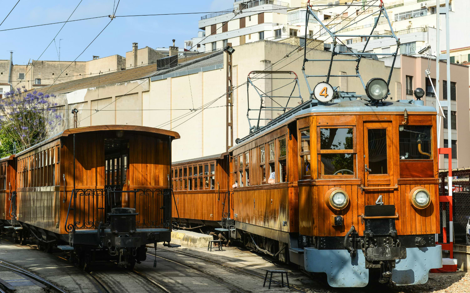 Vintage wooden carriages of the historic Sóller train stand at a station near Palma, framed by surrounding buildings and overhead wires.