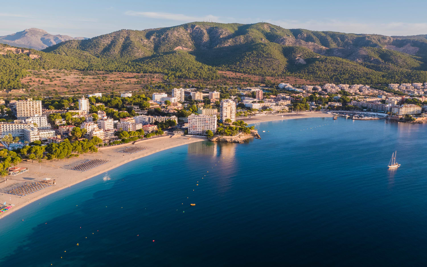 An aerial view of a coastal resort town near Palma de Mallorca, with a curved sandy beach, hotels and forested mountains behind.