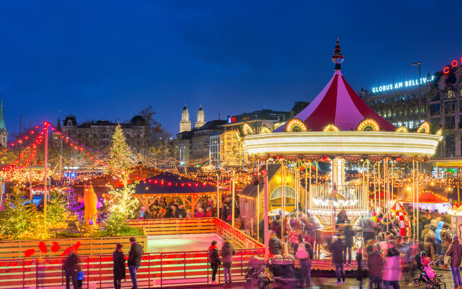 Christmas market at Zurichās Bellevue with a lit carousel, ice rink and Christmas trees, Grossmünster towers behind