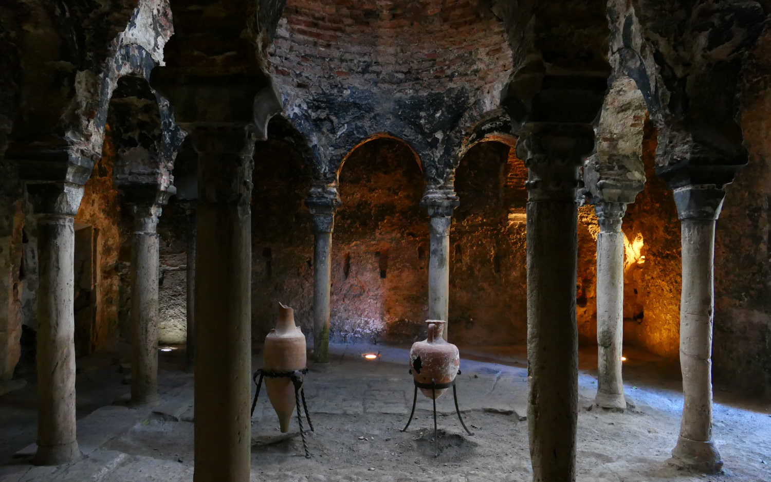 Horseshoe arches and weathered stone columns fill Palma's historic Arab Baths, with ancient clay amphorae resting on the sandy floor below