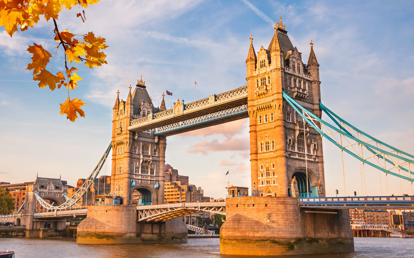 Tower Bridge glowing in warm evening light over the Thames, with yellow autumn leaves in the foreground