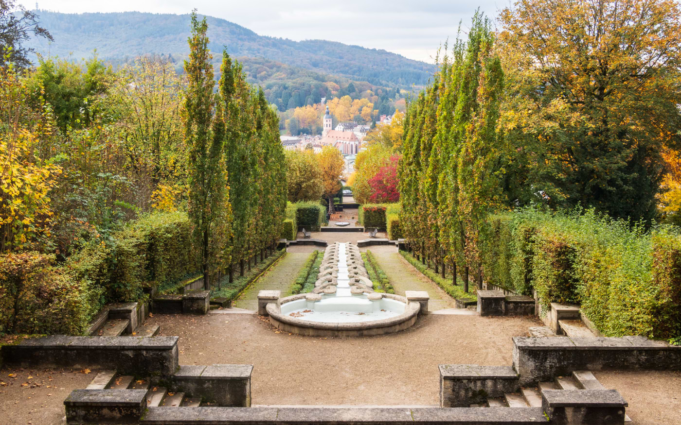 Terraced water cascade of the Paradies gardens in Baden-Baden, lined with tall trees, with autumn hills and a church behind