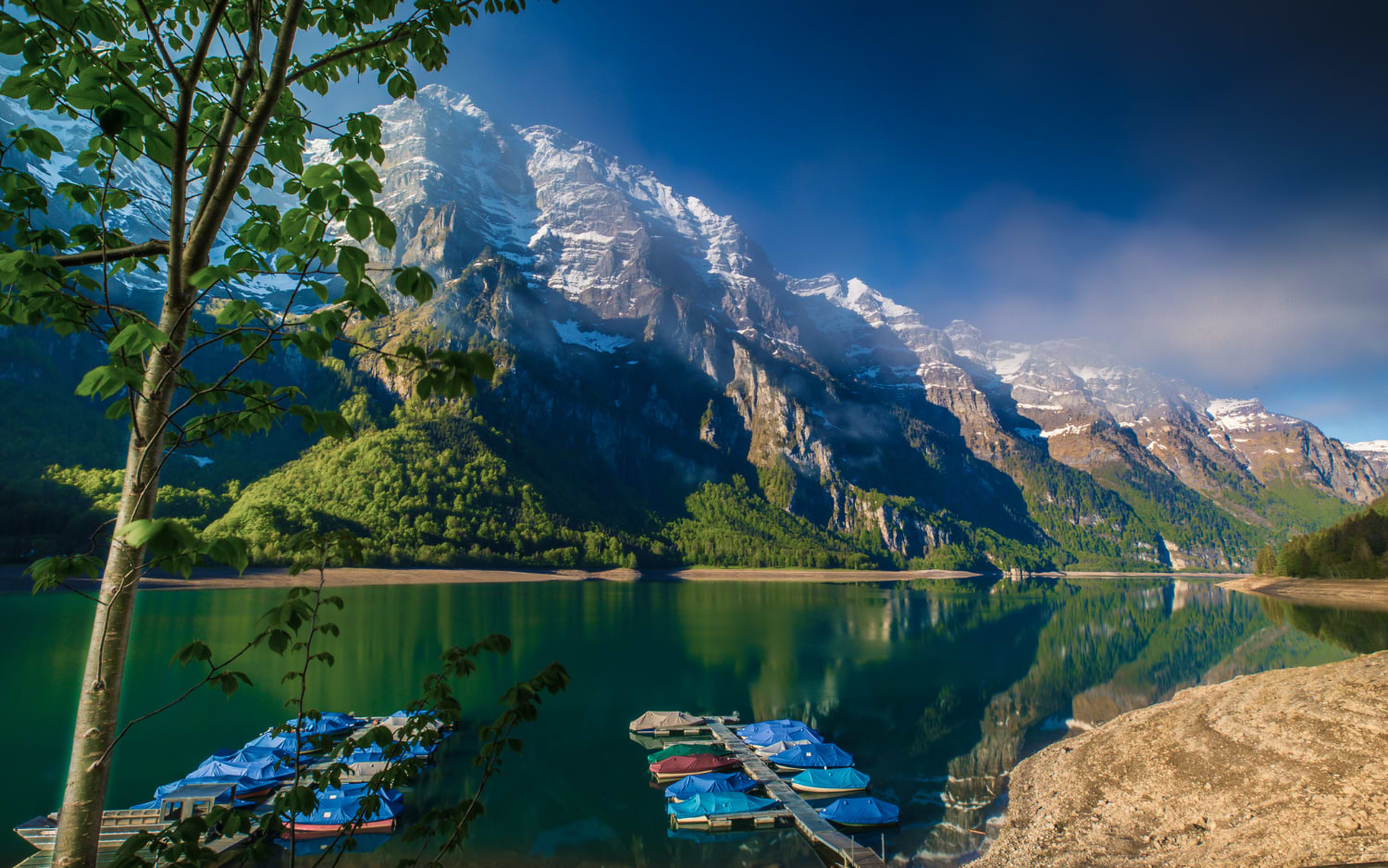 Turquoise alpine lake beneath steep, snow-streaked cliffs with small boats, like the Klƶntalersee walk