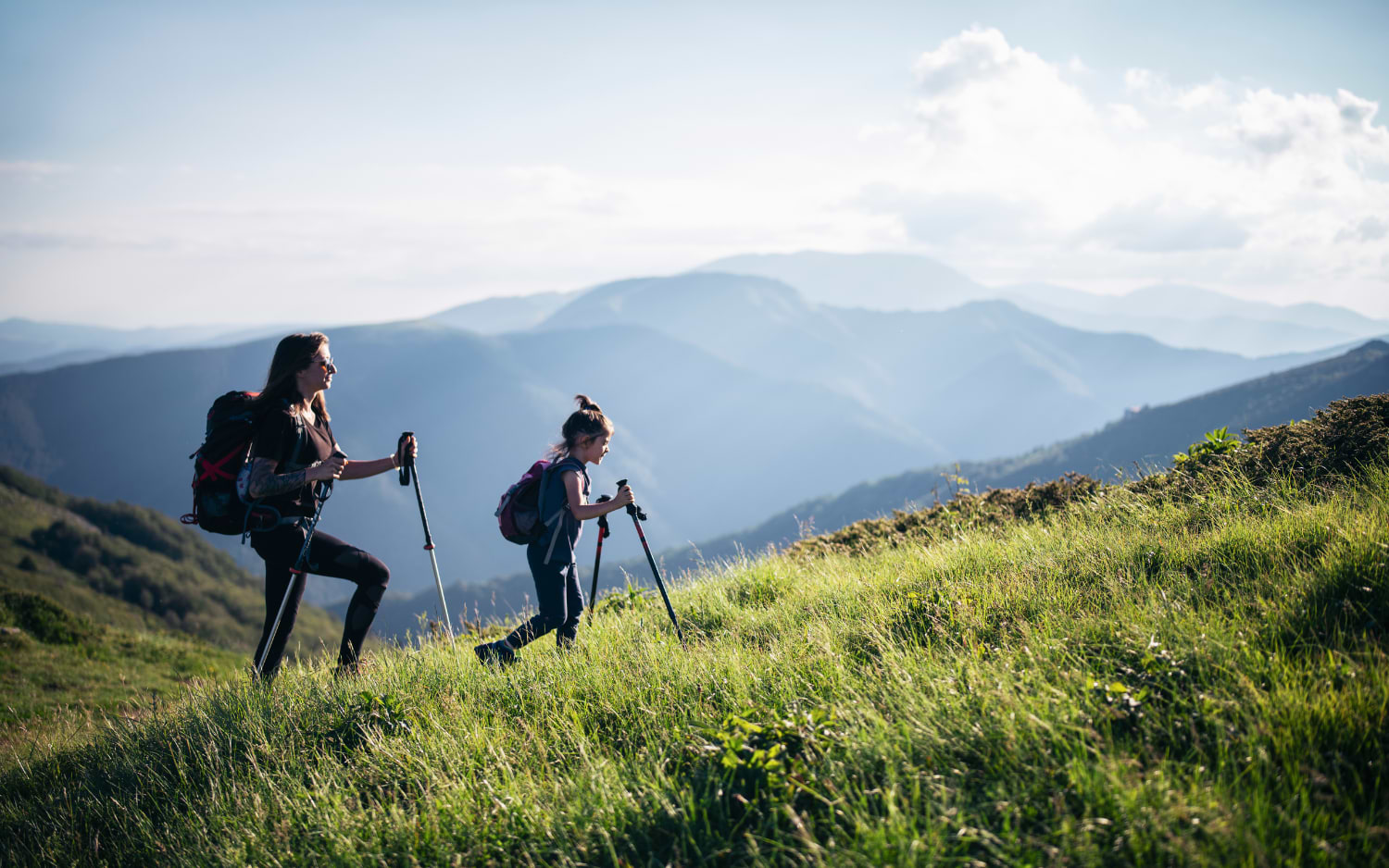 Two hikers with trekking poles climbing a grassy alpine slope, misty mountain ridges in the distance