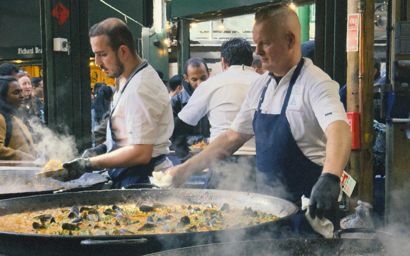 Chefs stirring giant steaming pans of seafood paella with mussels at a street food stall in Borough Market
