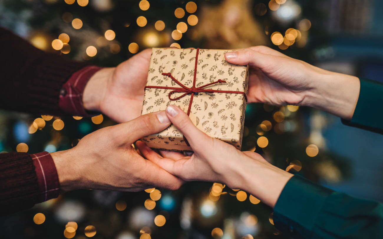 Zwei Paar Hände überreichen ein kleines Geschenk in gemustertem braunem Papier mit roter Schnur vor einem leuchtenden Weihnachtsbaum
