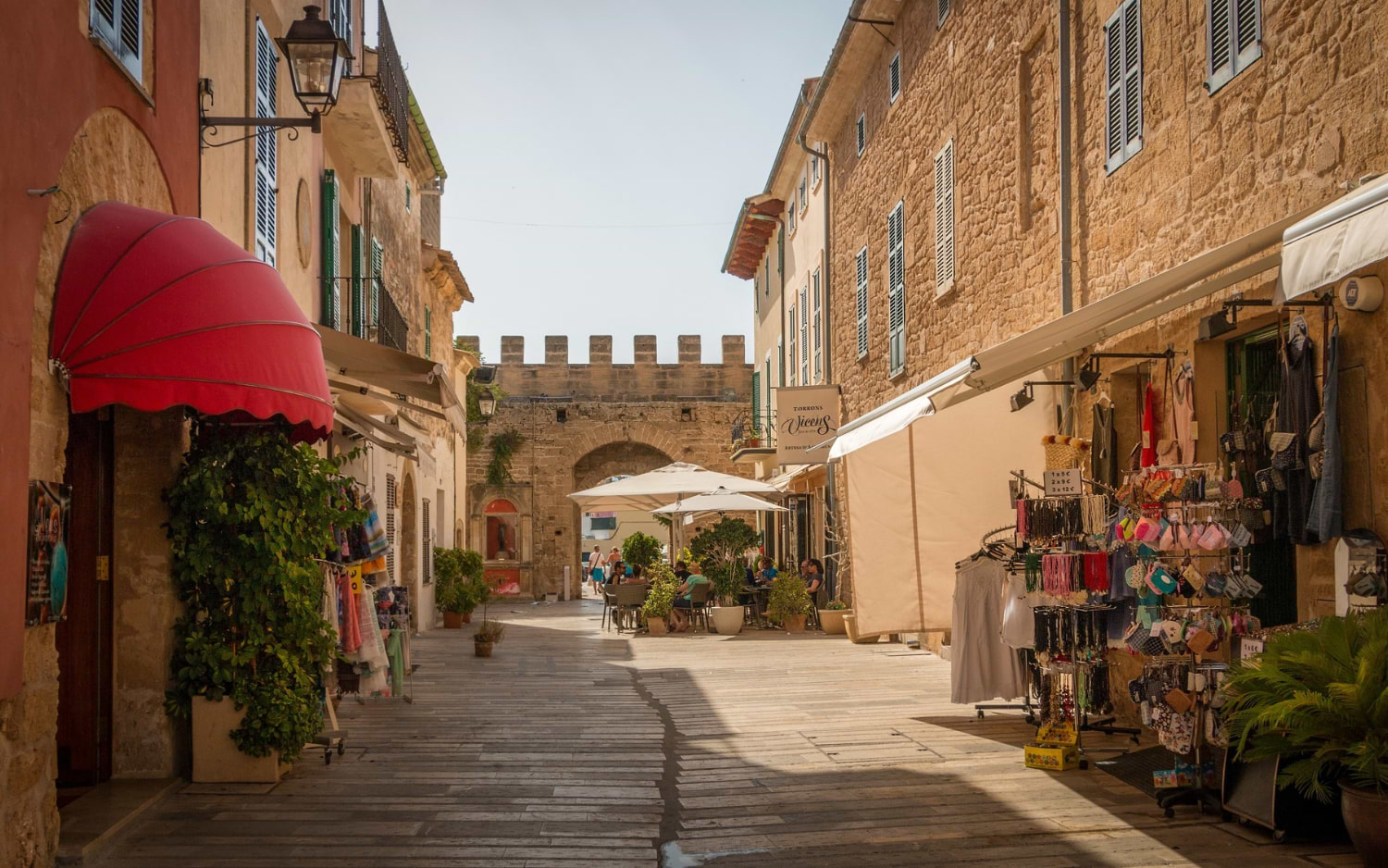 A sunlit cobbled street in AlcĆŗdia's old town near Palma, lined with souvenir stalls, cafes and a fortified gateway in the distance.