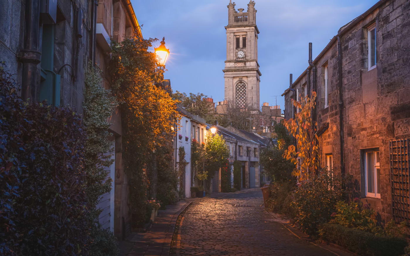 Cobbled Circus Lane in Edinburgh at dusk, lit by street lamps, with autumn leaves and the St Stephen's Church clock tower