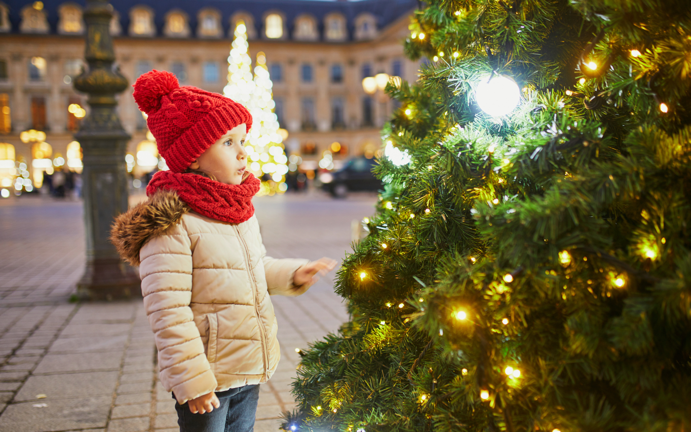 Child in a red knitted hat and scarf gazes at a lit Christmas tree on a festive Paris square during the Christmas season