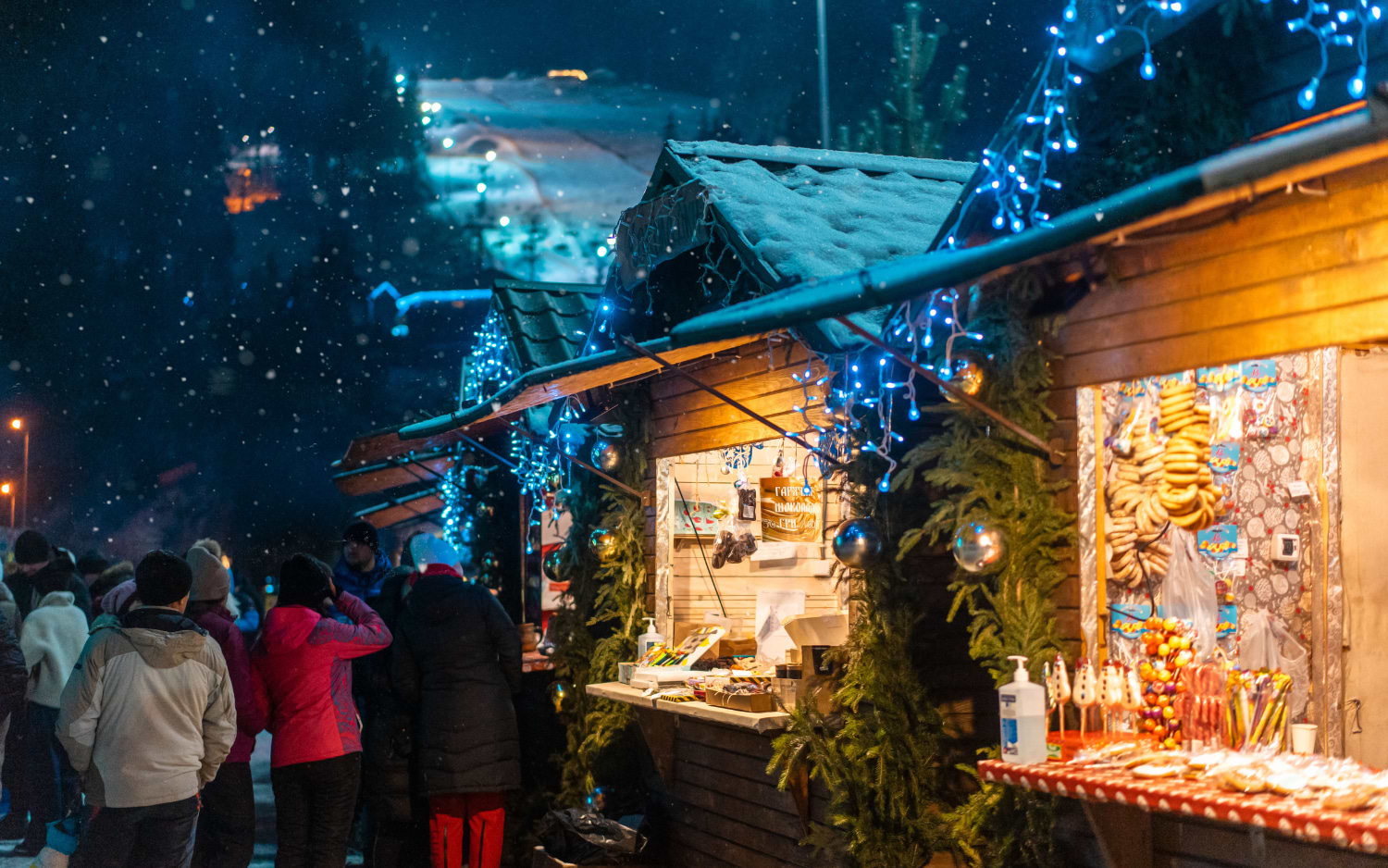 Snow falls on lit wooden Christmas market stalls selling sweets and pastries, with a floodlit ski slope behind
