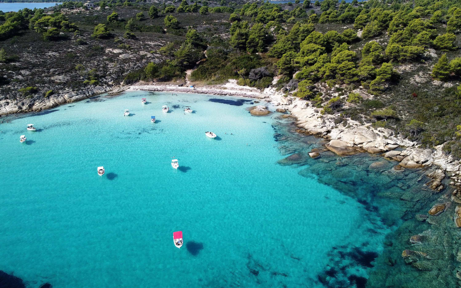 Aerial view of a secluded turquoise bay in Halkidiki with small boats and pine-covered rocky shores
