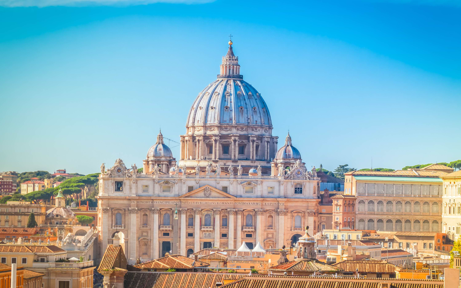 Luftaufnahme der Kuppel des Petersdoms über Roms Ziegeldächern vor blauem Himmel