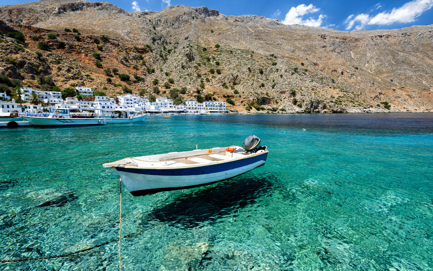 Small blue-and-white motorboat moored in clear turquoise water, Crete village and rocky mountain behind
