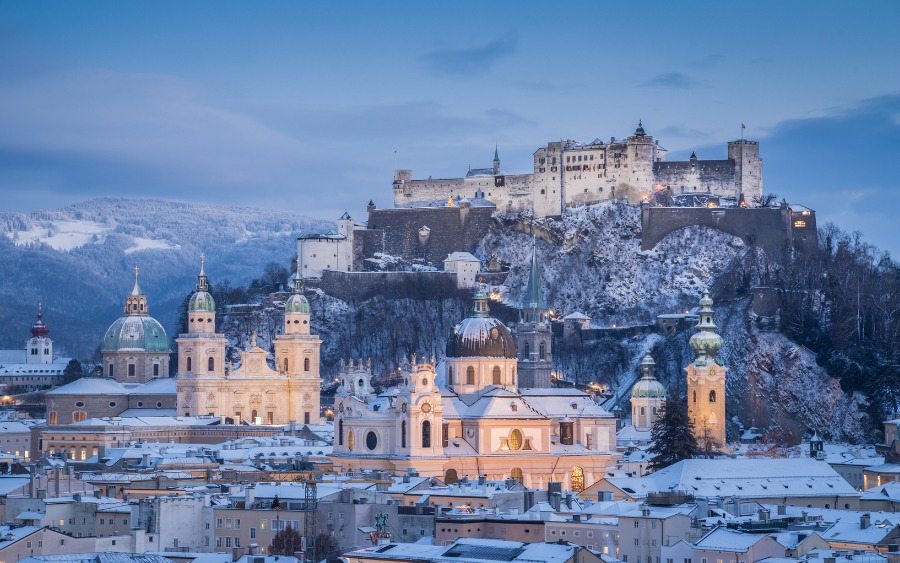 A panoramic view of Salzburg, Austria, showcasing snow-dusted rooftops and the Hohensalzburg Fortress under a clear winter sky