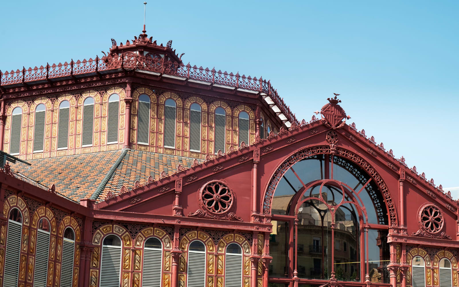 Ornate red ironwork facade and glass arches of a historic Barcelona market building under blue sky.