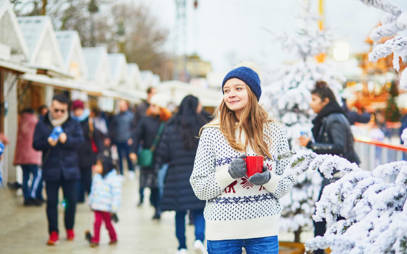 Young woman in a Nordic sweater holds a red mug of mulled wine by white chalets and snowy trees at a Paris Christmas market