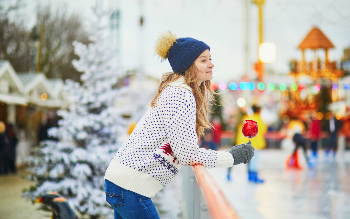 Woman in a blue bobble hat holds a red candy apple at an ice rink railing, with Christmas market fairground lights behind