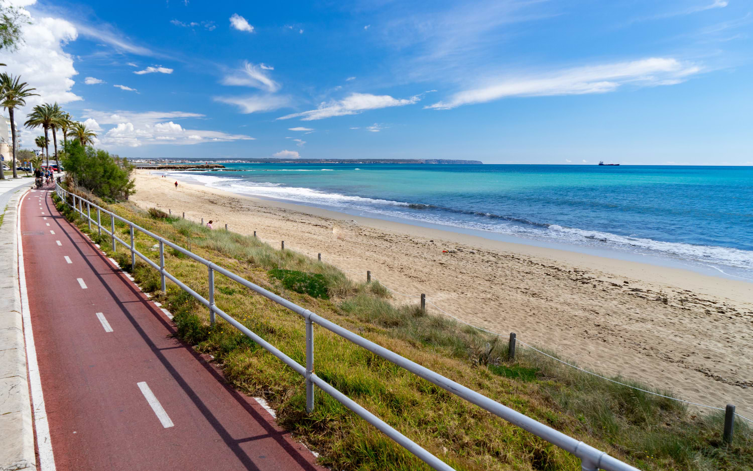 A red cycling path runs alongside the sandy shore of Playa de Palma, with palm trees, turquoise sea and distant cliffs beyond the bay