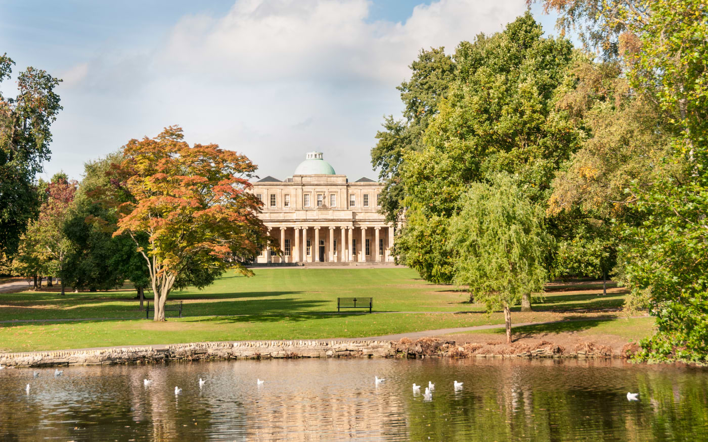 Pittville Pump Room in Cheltenham, a domed Regency building behind a lake with gulls and trees turning orange in autumn