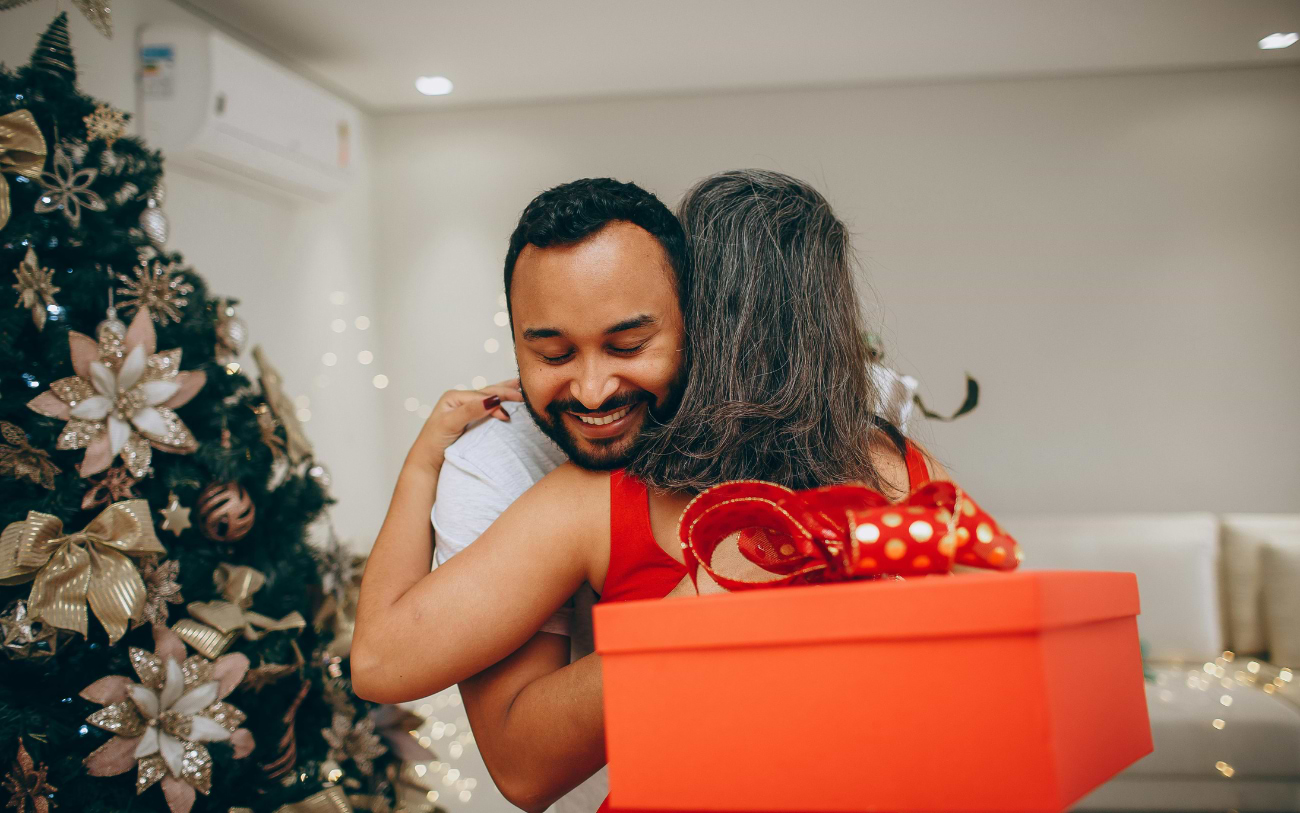 Ein lächelnder bärtiger Mann umarmt eine grauhaarige Frau mit großer roter Geschenkbox und gepunkteter Schleife neben dem Weihnachtsbaum