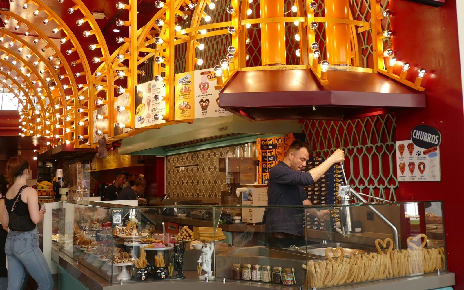 A vendor prepares fresh churros beside a glowing gelato and pastry counter inside the Time Out Barcelona market.