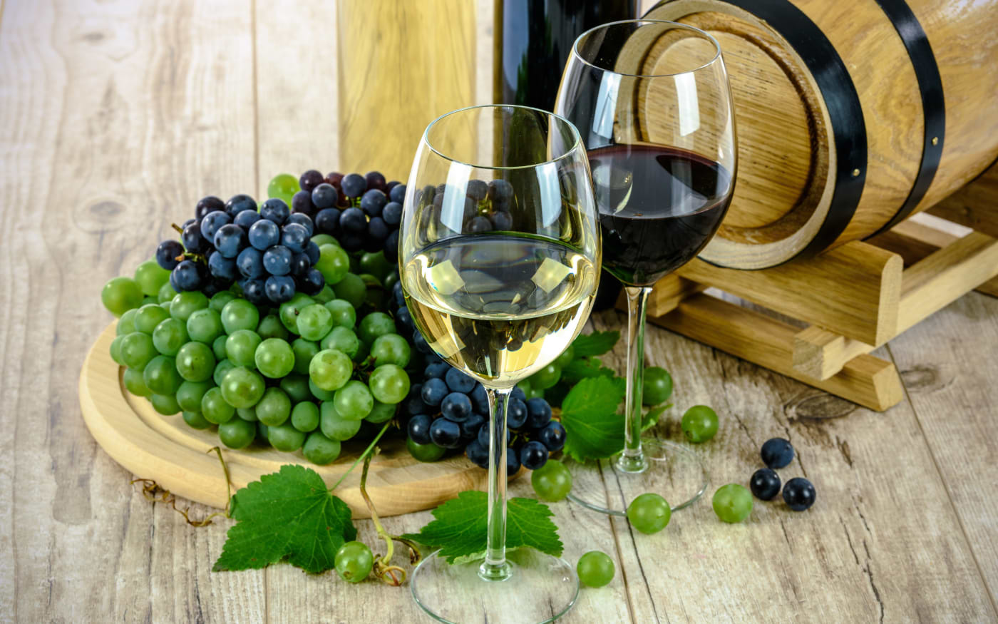Glasses of white and red wine beside grapes and a wooden wine barrel