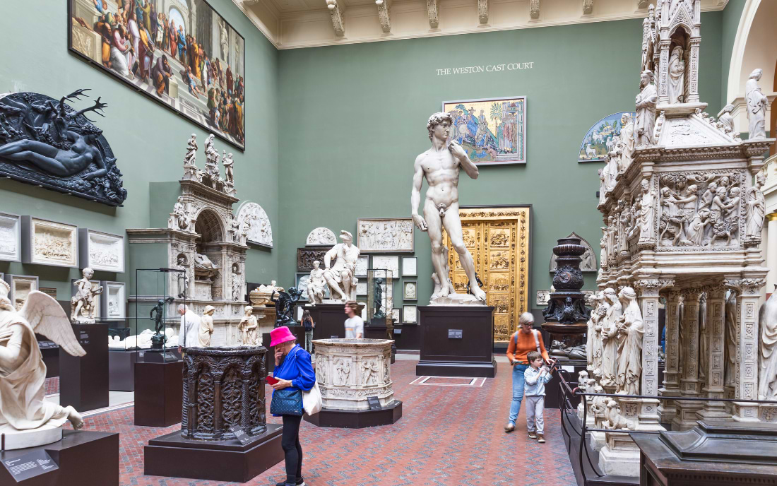 The Weston Cast Court at the Victoria and Albert Museum London, filled with large-scale plaster casts of European sculptures including a replica of Michelangelo's David, as visitors explore the green-walled gallery