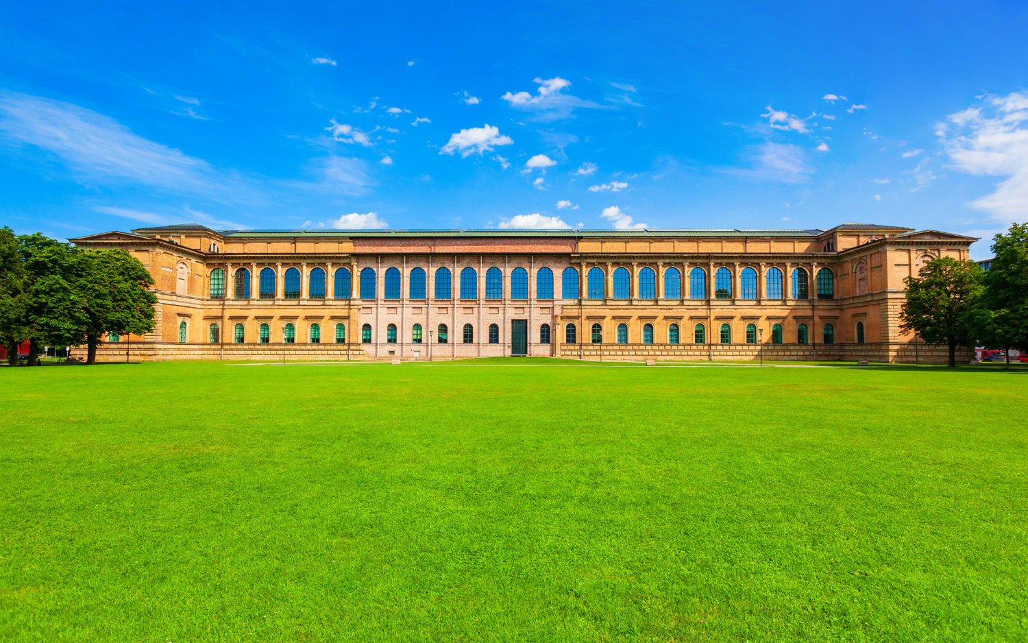 The Alte Pinakothek art museum in Munich, a long sandstone building with rows of arched windows, seen across its wide green front lawn.