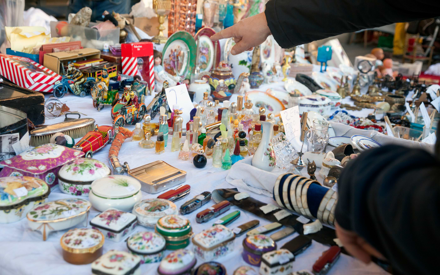 Buyer points to porcelain trinket boxes, vintage perfume bottles, tin toys and pocket knives at a Vienna flea market stall.
