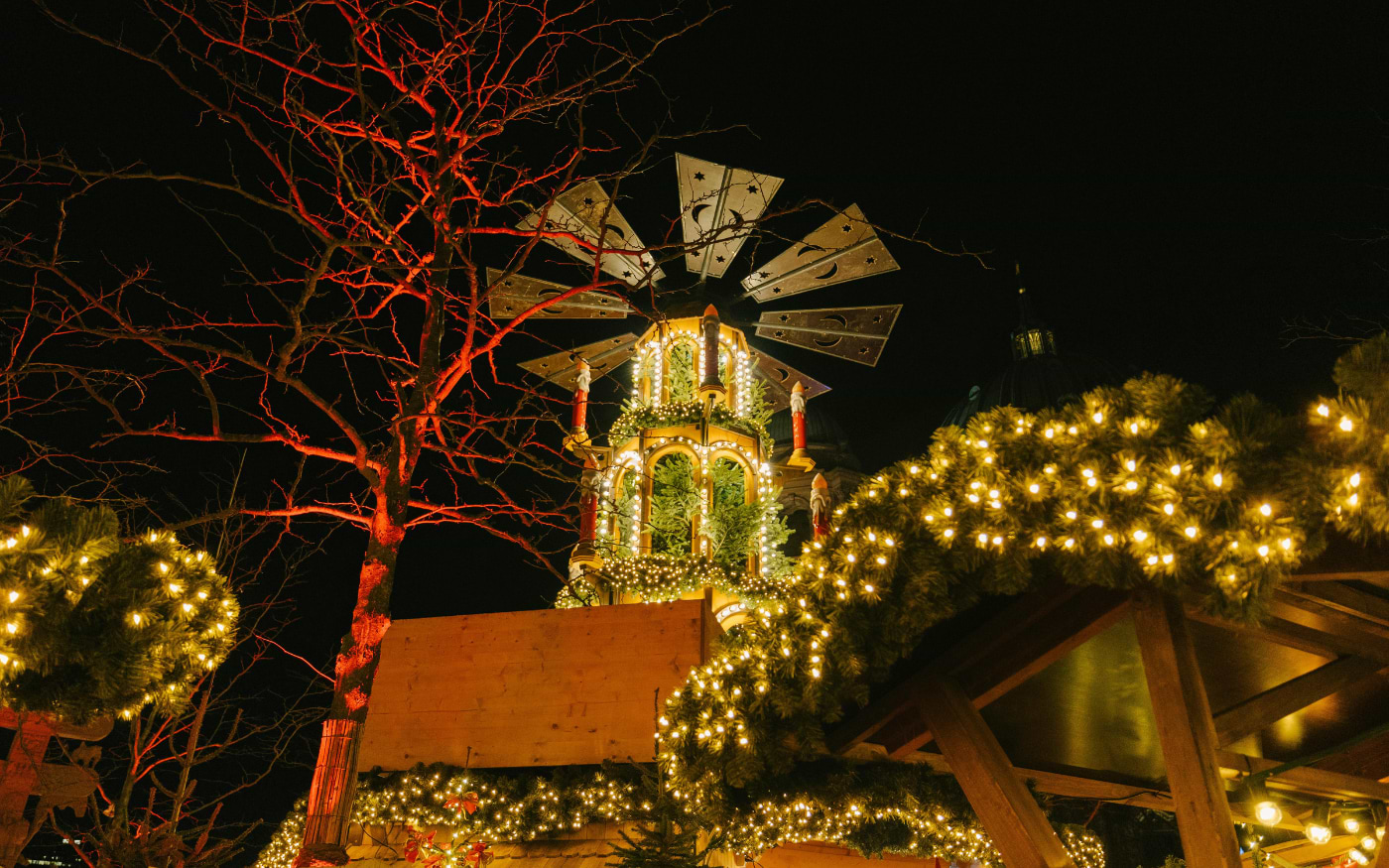 Giant wooden Christmas pyramid glowing with fairy lights beside a red-lit tree at a Berlin Christmas market at night