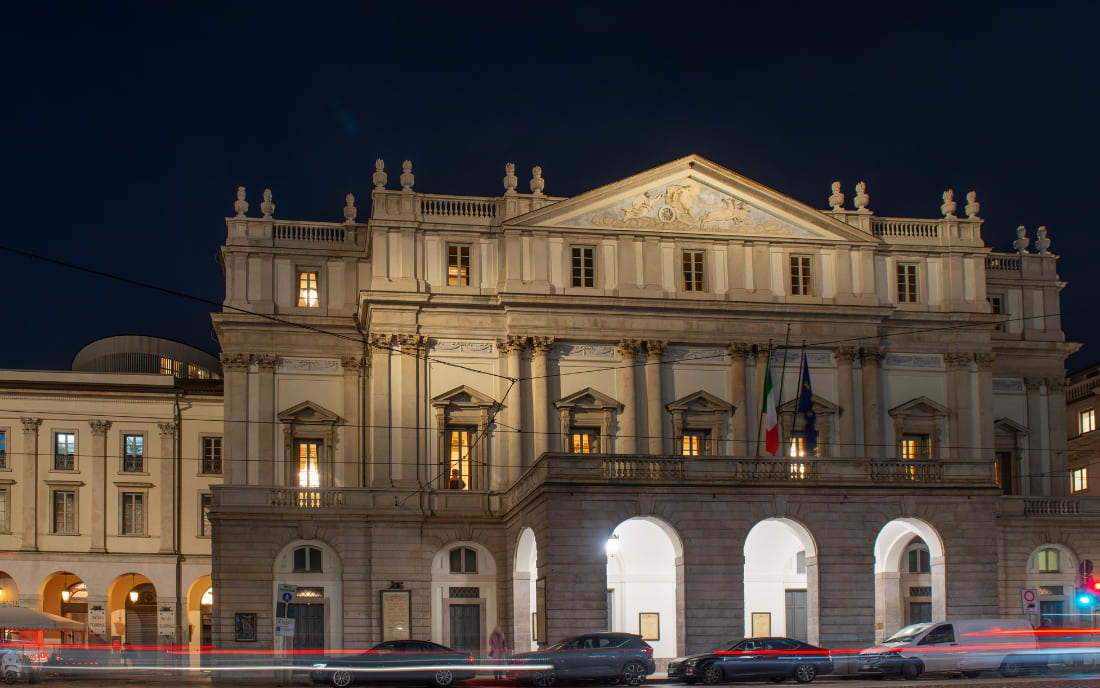 The illuminated neoclassical facade of Teatro alla Scala opera house in Milan at night, with light trails from passing traffic