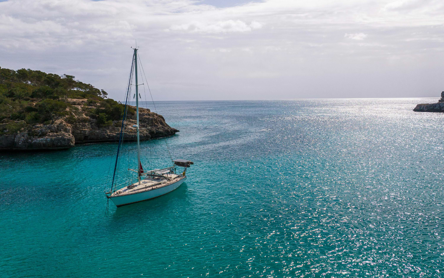 A white sailboat anchors in a turquoise cove near Palma de Mallorca, framed by pine-covered rocky headlands under a soft cloudy sky.