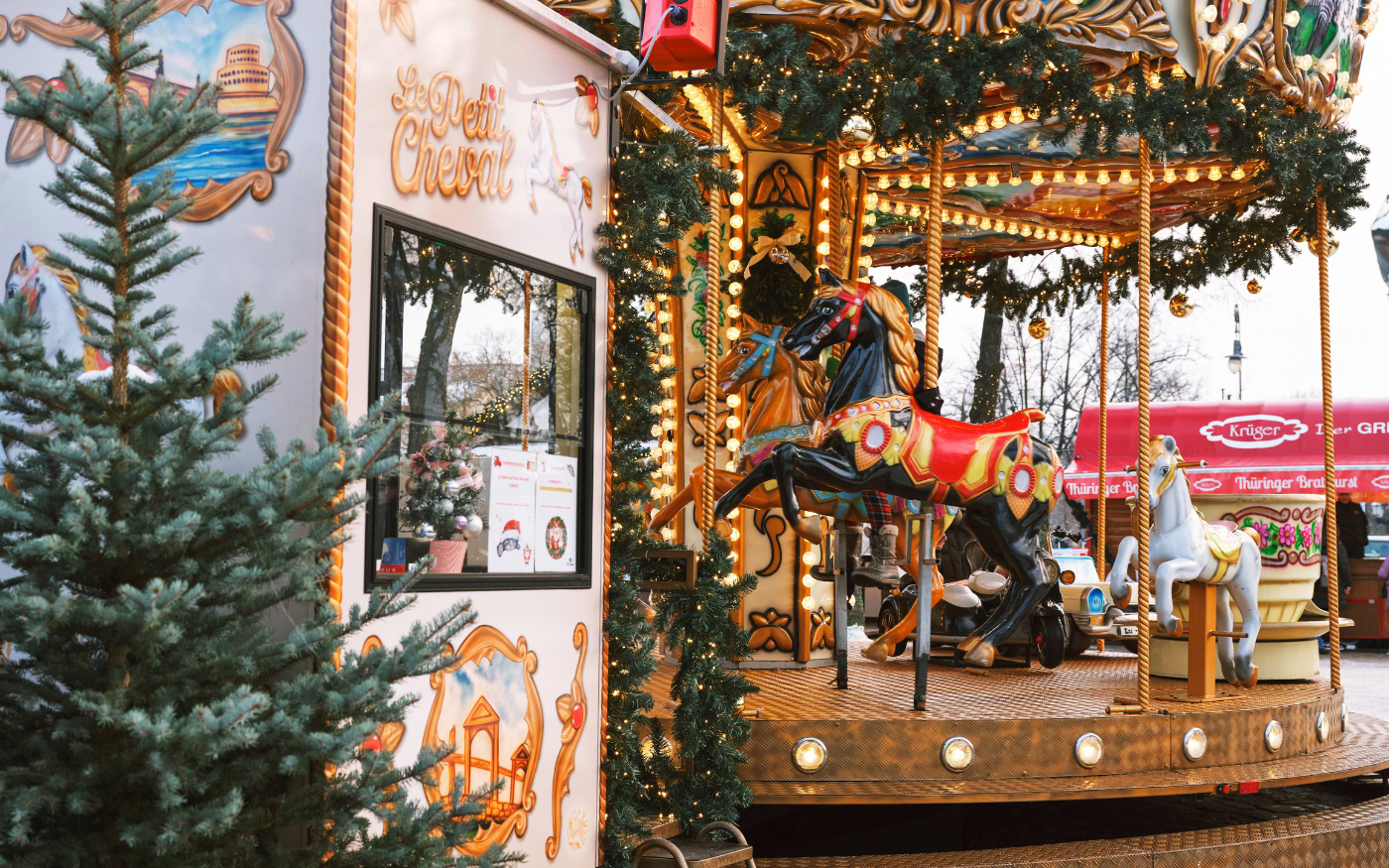 Vintage carousel with painted horses, gold lights and fir garlands beside the Le Petit Cheval booth at a Christmas market