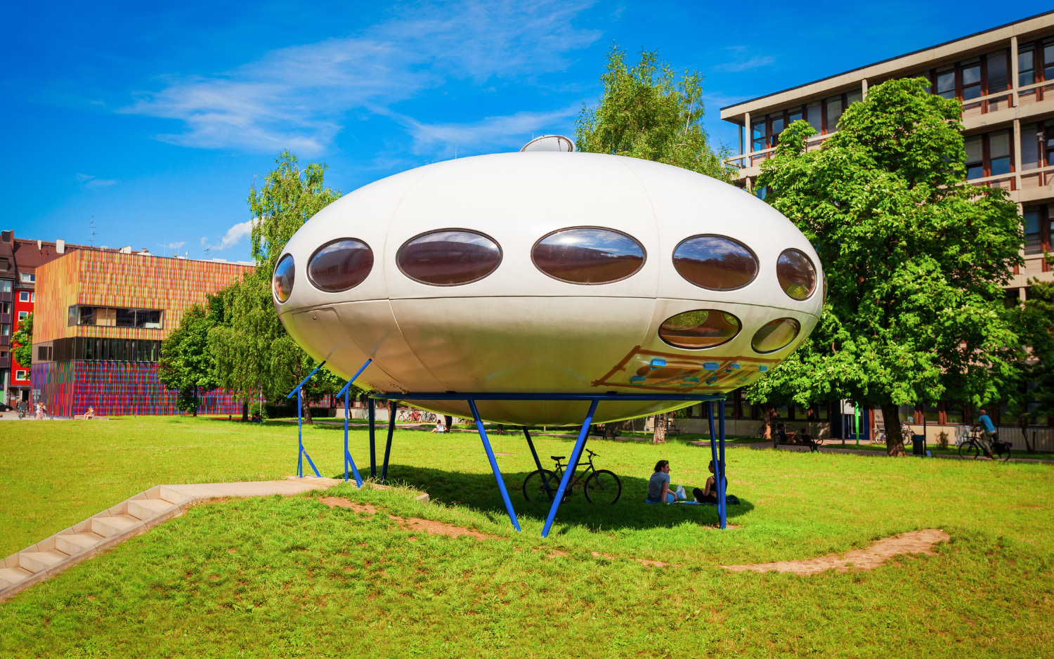 A retro white Futuro House, an oval fibreglass pod raised on blue steel stilts, standing on a lawn in Munich's Kunstareal district.