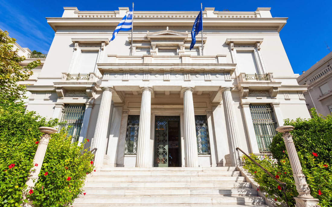 Exterior facade of the Benaki Museum Athens, a white neoclassical mansion with Greek and EU flags, surrounded by lush greenery