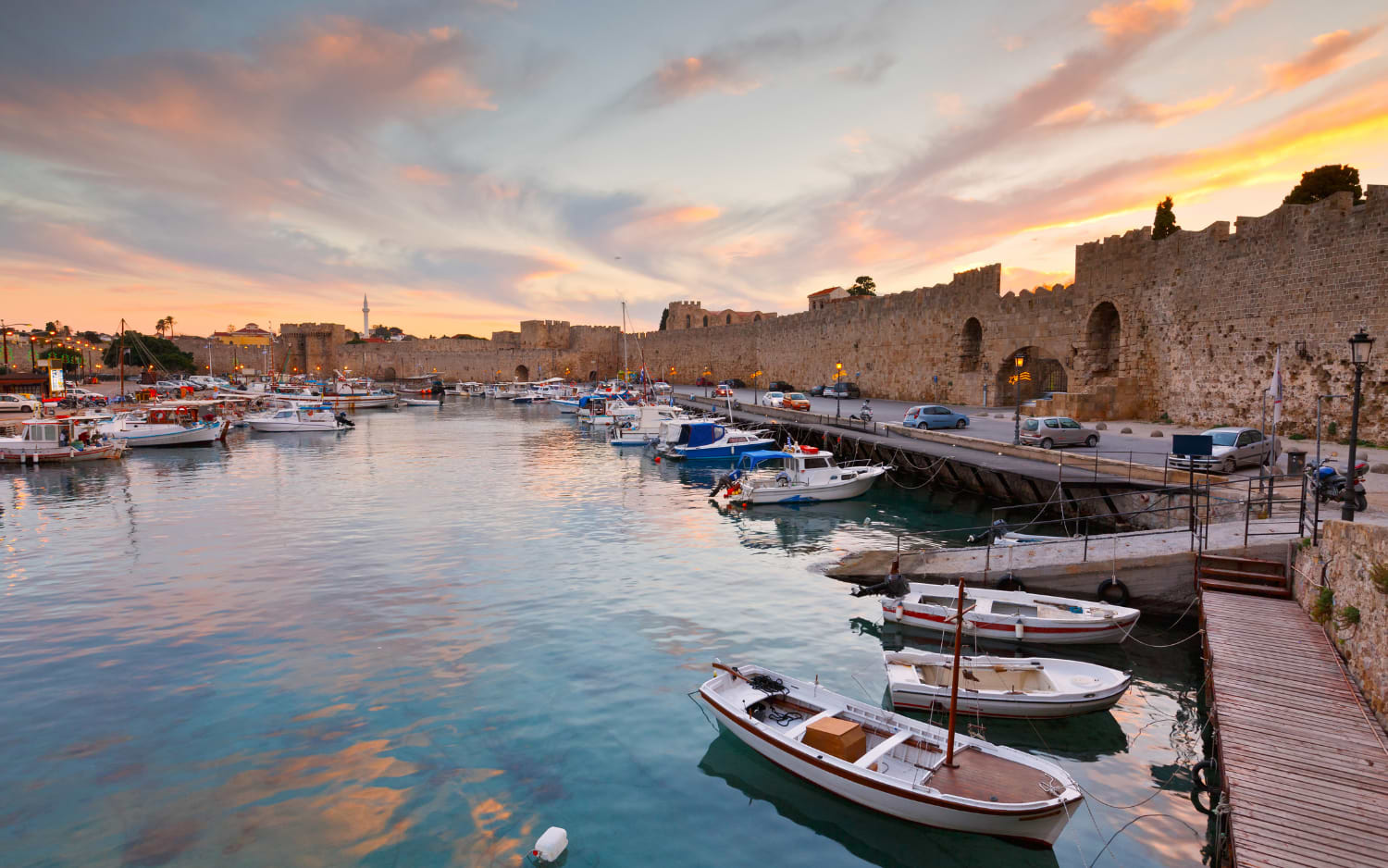 Rhodes Old Town harbour at sunset with fishing boats and medieval city walls glowing in orange light