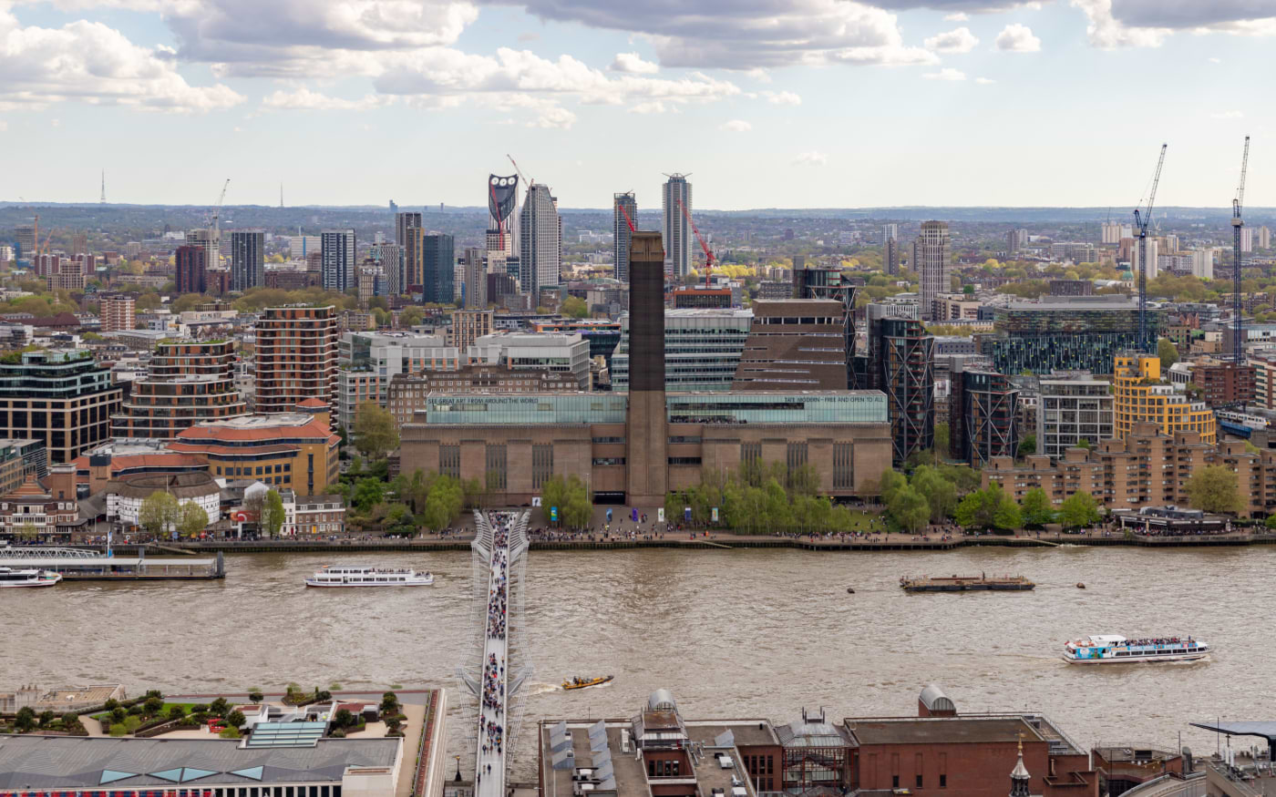 Aerial view of Tate Modernās brick chimney and the crowded Millennium Bridge crossing the Thames, with tour boats below