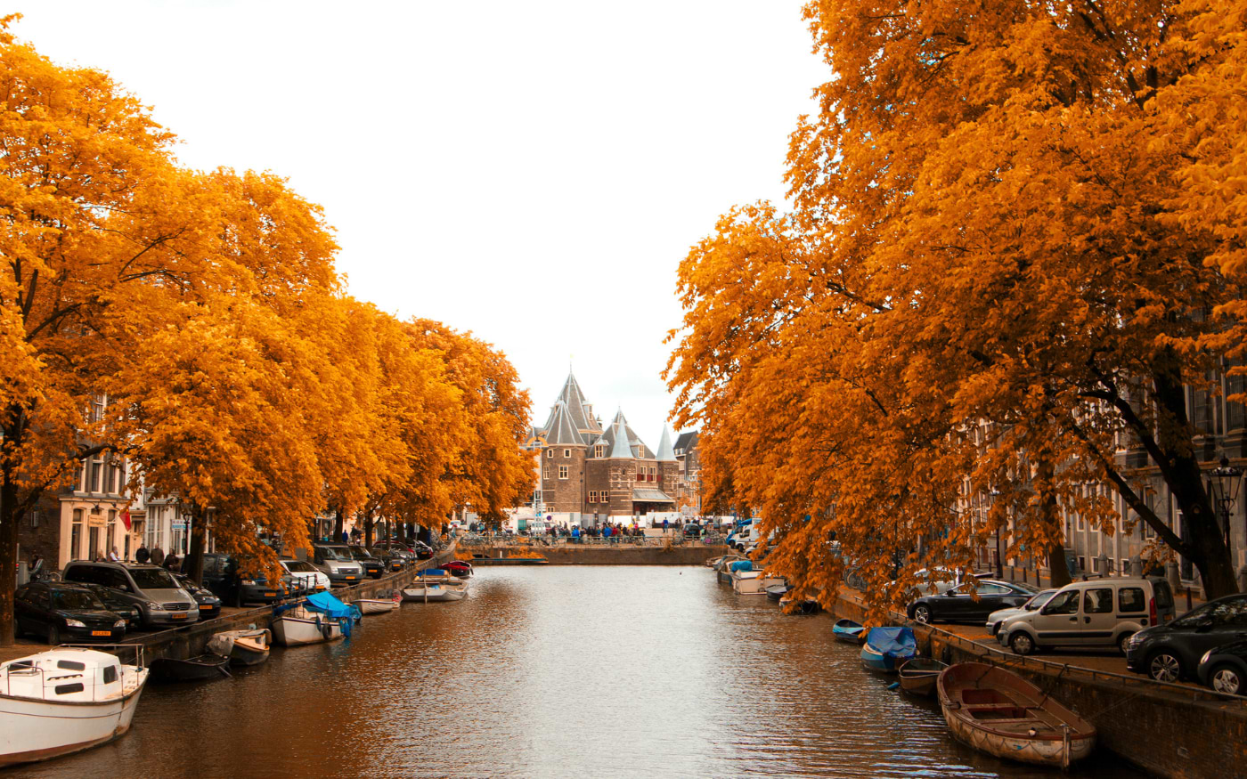 Amsterdam canal lined with bright orange autumn trees and moored boats, leading to the turreted De Waag building