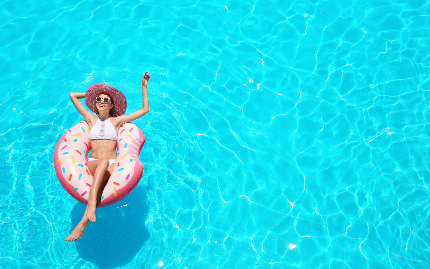 Woman relaxing on a donut inflatable in a turquoise pool, sun hat and sunglasses, overhead view