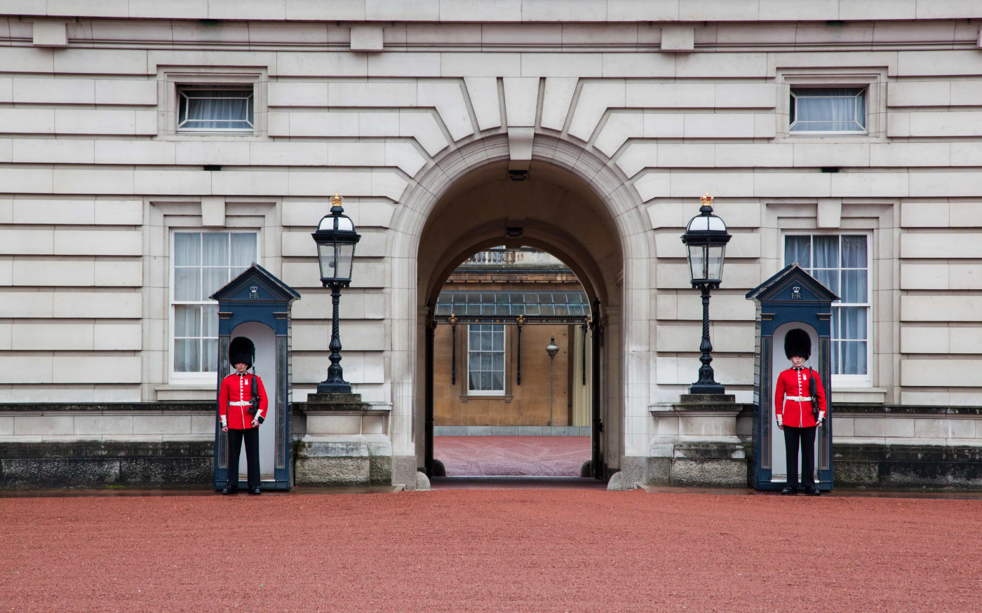 Two guards in red tunics and bearskin hats beside sentry boxes at a Buckingham Palace archway, free to see in London
