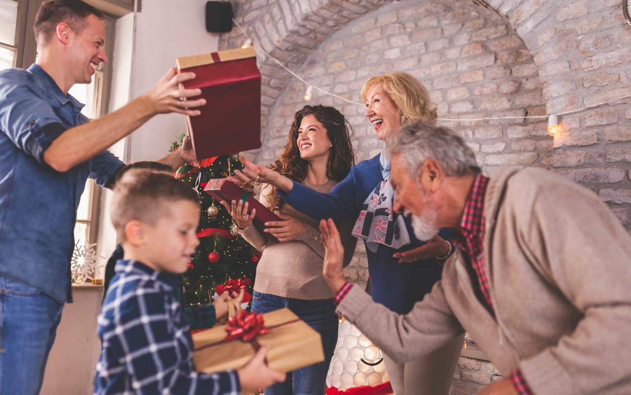 Drei Generationen einer Familie lachen und reichen sich verpackte Geschenke in einem festlichen Raum mit Backsteinwänden und Lichterketten