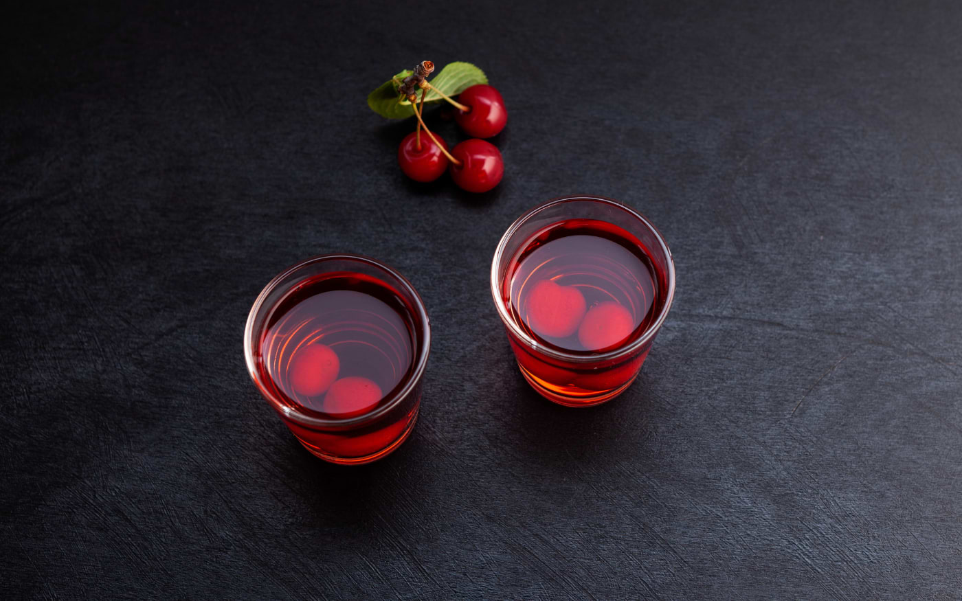 Two glasses of ginjinha, traditional Portuguese cherry liqueur, served with fresh cherries on a dark table.