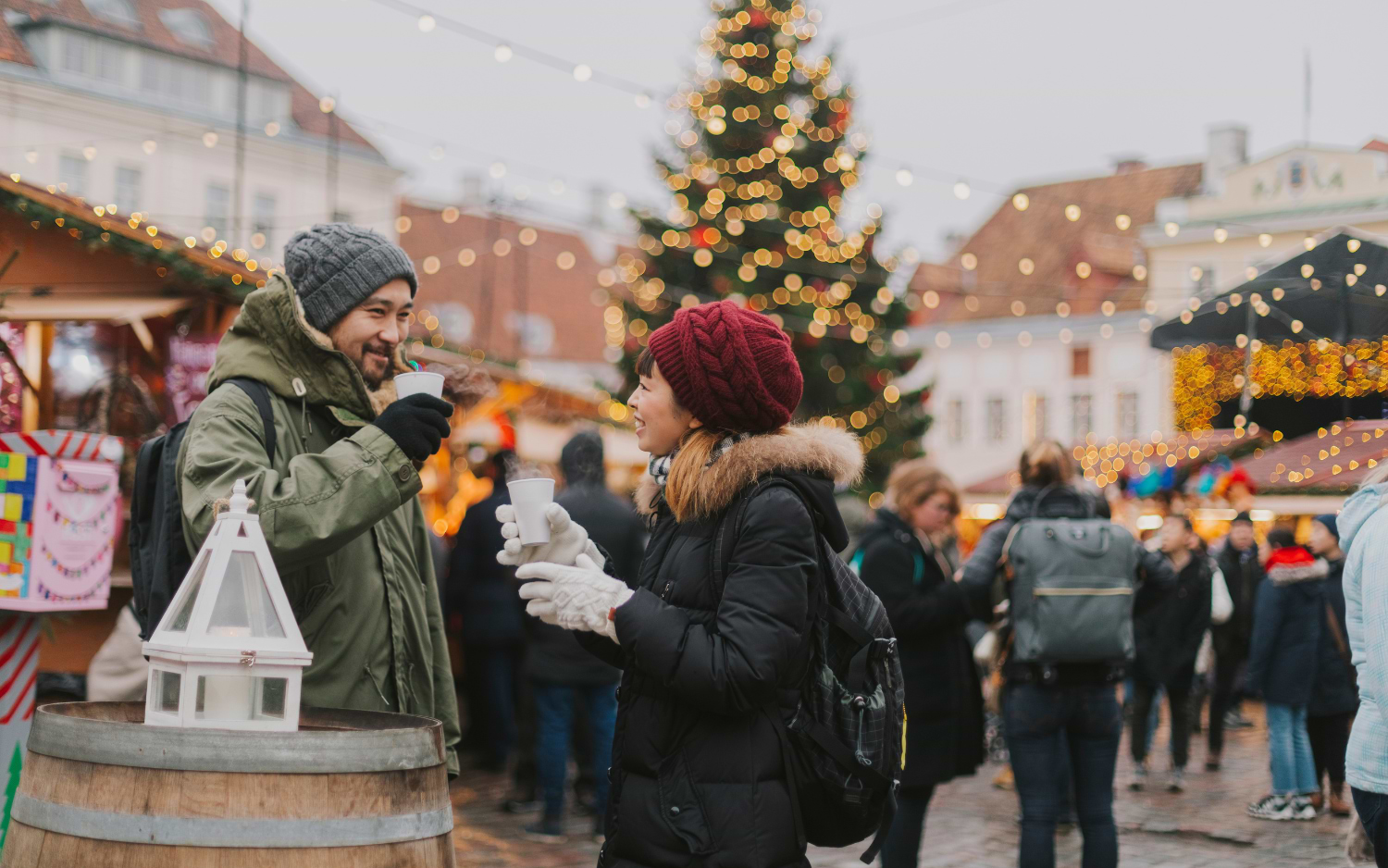 Smiling couple in hats toasting with mulled wine cups at a Christmas market, a lit tree and stalls behind