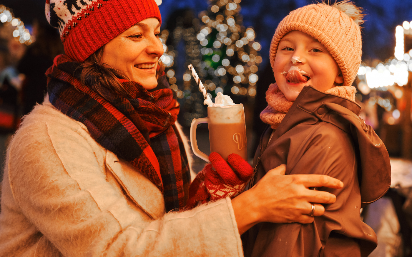 Smiling mother and child, with cream on the child's nose, sharing hot chocolate at a Berlin Christmas market at night