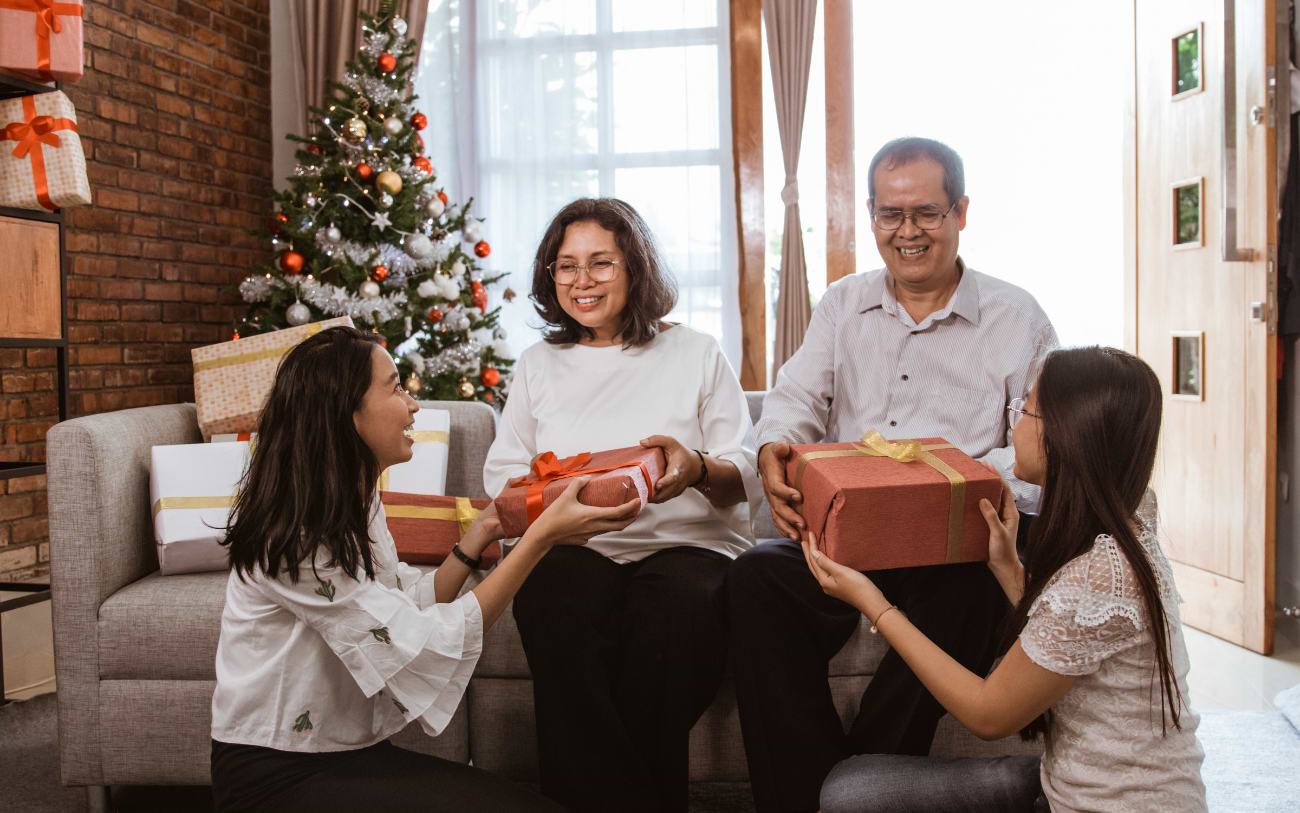 Zwei junge Frauen überreichen einem älteren Paar auf dem grauen Sofa Geschenke mit orangefarbenen Schleifen neben dem Weihnachtsbaum