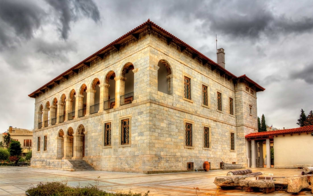 Exterior of the Byzantine and Christian Museum Athens, a two-storey stone building with arched colonnades and a terracotta roof under a dramatic cloudy sky